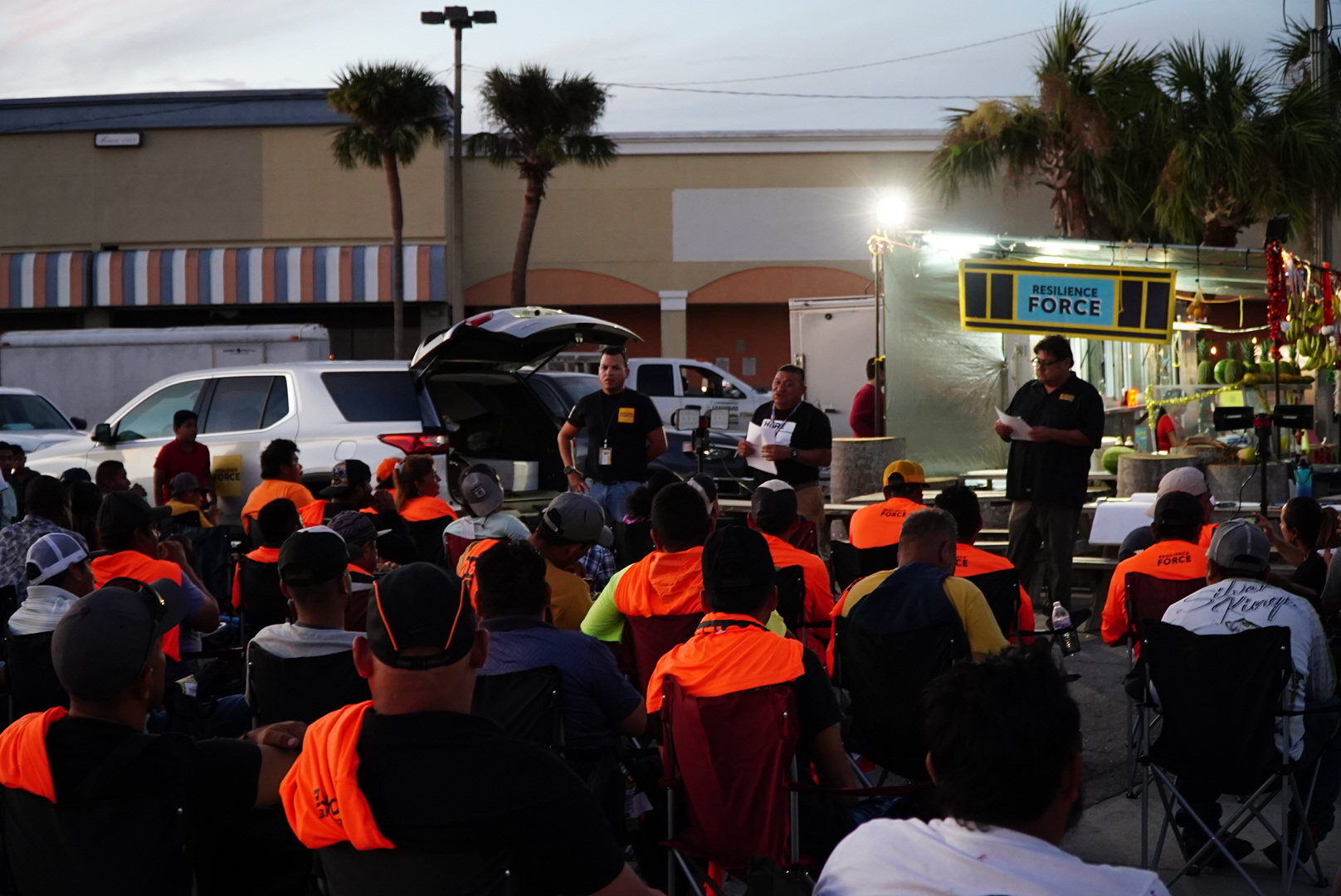 Resilience Force activist and former disaster restoration worker, Mariano, stands in the middle of his two co-workers holding documents while speaking to workers in November 2022, two months after Hurricane Ian devastated the region. Scores of disaster restoration workers, some dressed in bright orange long sleeve shirts, listen during the first worker meeting organized by Resilience Force in Fort Myers, post-Ian. (Jiahui Huang/Columbia Journalism Investigations)